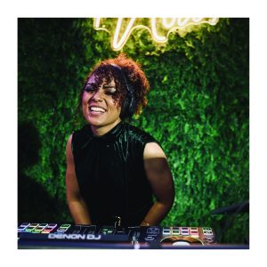 DJane von Music Nau Now lächelt hinter dem DJ-Pult mit Kopfhörern bei einem hochwertigen Event.