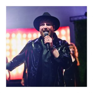 Sänger mit Hut und Mikrofon bei einem energiegeladenen Live-Auftritt von Music Nau Now auf einem exklusiven Event.