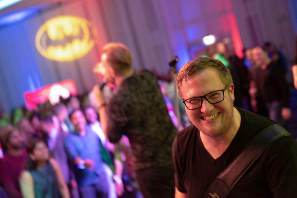 Gitarrist von Music Nau Now lächelt in die Kamera während eines Live-Showacts vor feierndem Publikum.