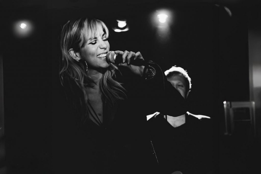 Schwarzweißaufnahme einer Sängerin am Mikrofon während eines Live-Auftritts, emotionaler Ausdruck, Bühnenlicht im Hintergrund