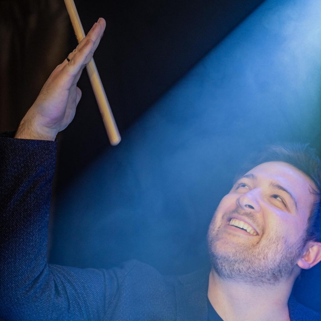 Schlagzeuger und Percussionist von Music Nau Now hebt den Drumstick im blauen Bühnenlicht und dirigiert den nächsten Break während der Liveband-Performance